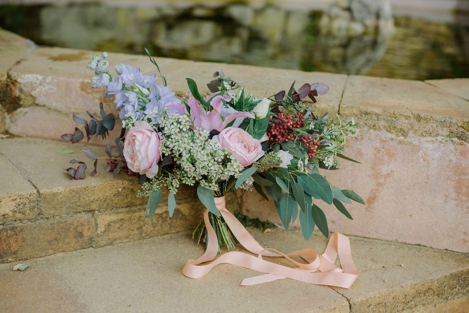 A delicate floral bouquet arranged with soft pink roses, pale purple delphiniums, white astilbes, and sprigs of red berries, complemented by green eucalyptus leaves and stems, all tied together with a