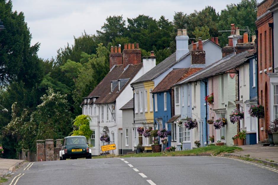 A quiet residential street in Berkhamsted, featuring a row of colorful terraced houses painted in pastel shades of white, yellow, blue, and light green. Each house has a small front garden with vibran