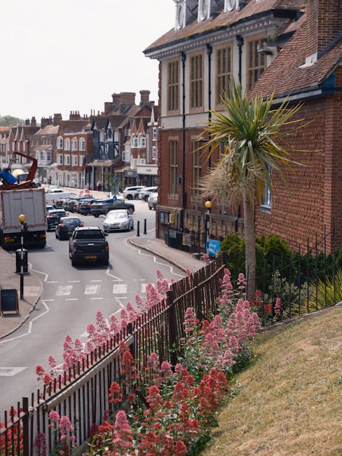 A view of a residential street in Berkhamsted featuring a flower bed with pink and red flowering plants along a black wrought iron fence, a tall palm tree with long, spiky green and brown leaves stand