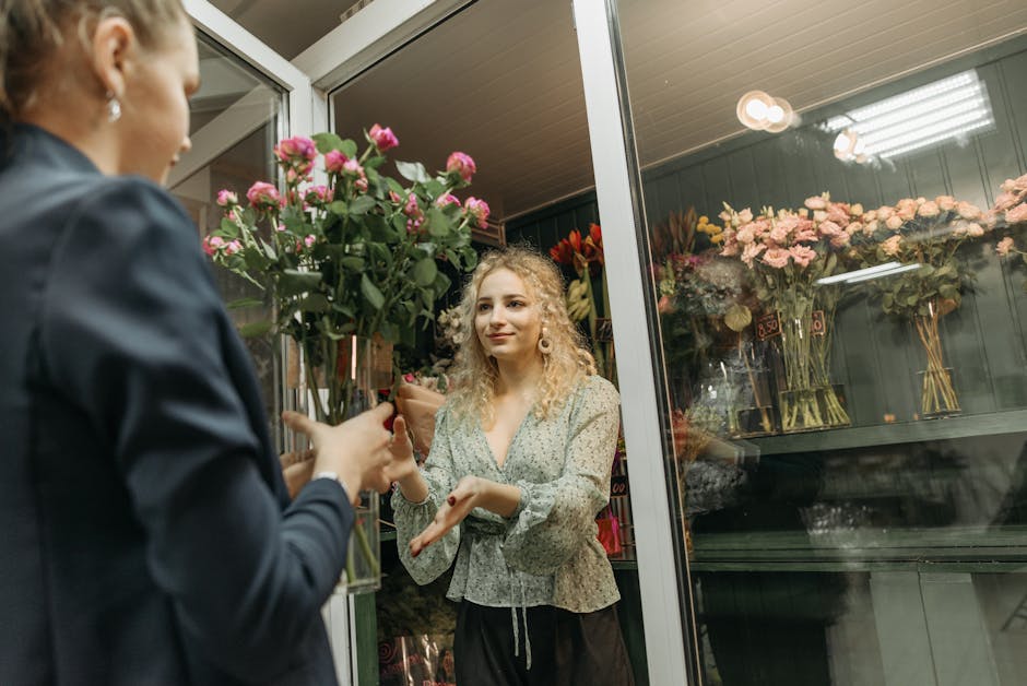 A woman with curly blonde hair, wearing a light-colored floral blouse, stands inside a flower shop at night, smiling and gesturing towards a large, vibrant bouquet of pink roses with green foliage, wh