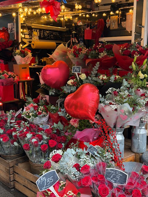 A vibrant display of floral arrangements at a flower stall, featuring predominantly red roses and white baby's breath, wrapped in pink and red paper with some accented by green foliage. Large red hear